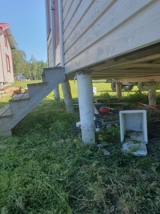 Husgrund stöttad av provisoriska pelare, gräsmatta, trädgårdsutrustning, lantlig miljö, klart väder, dagsljus.