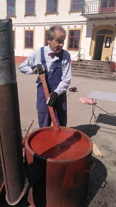 Man i blåkläder omrör röd vätska i Tunna framför historisk byggnad, utomhusarbete, traditionellt hantverk, solig dag.