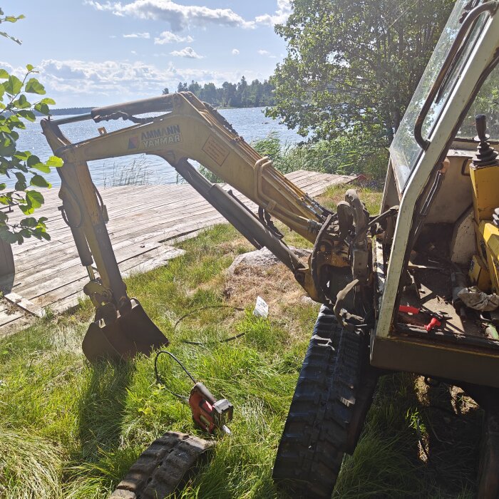 Grävmaskin nära sjö med skadad hytt, bredvid träbrygga, soligt, grönska.