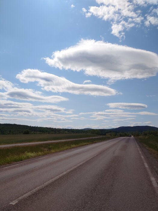 En öde landsväg sträcker sig rakt fram, omgiven av öppna fält, skogar och dramatisk himmel med ovanliga molnformationer.