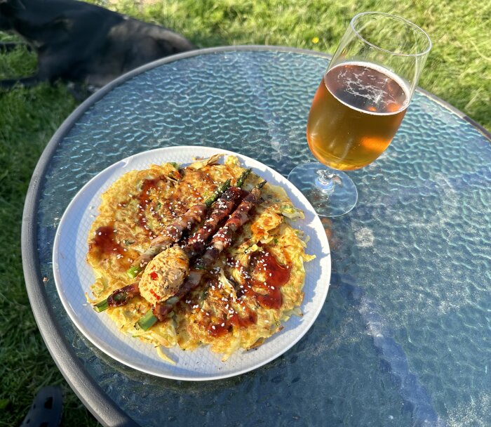 En tallrik med japansk okonomiyaki och en öl på ett glasbord utomhus. Skymningen nära, hund i bakgrunden.