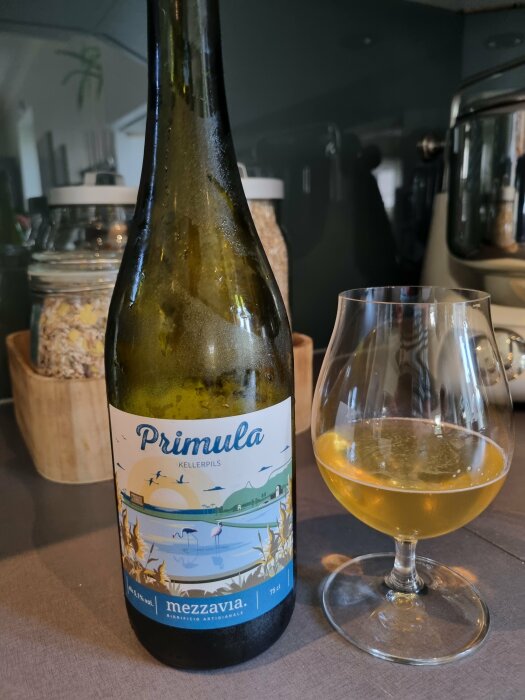 Flaska märkt "Primula Kellerpils", färgad dryck i glas, inomhusmiljö, köksomgivning.