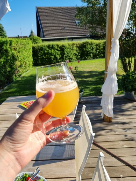 En hand håller ett ölglas på solig veranda med grönt gräs och delvis skymd sikt av hus.