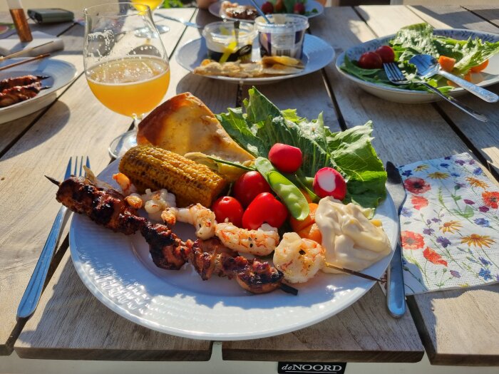 Middag utomhus med grillad mat och öl i solljus på ett träbord.