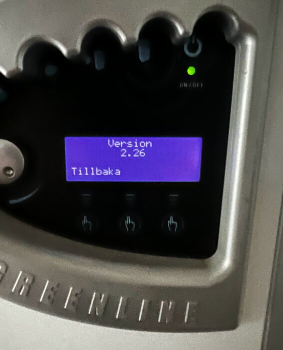 Elektronisk enhet med blått LCD-gränssnitt visar "Version 2.26" och texten "Tillbaka", omgiven av knappar.