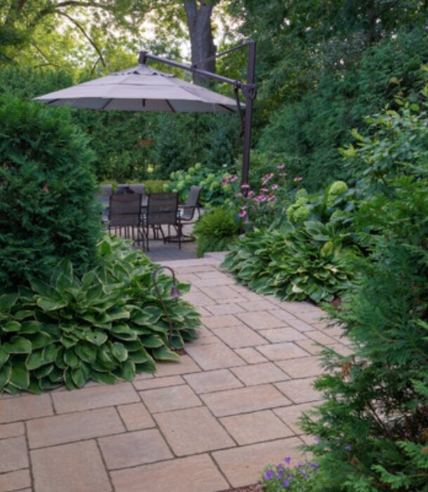 Ett mysigt trädgårdsutrymme med stenstig, uteplats, parasoll och grönskande växtlighet runtomkring.