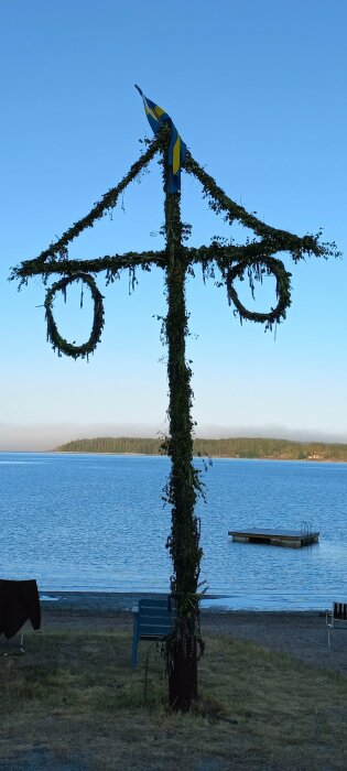 Svensk midsommarstång vid sjö, blå himmel, svenska flaggan i topp, brygga, bänk och badplats syns.