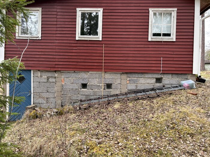 Rödfärgat hus, sockel av gråa betongblock, nedfallna saker, två fönster, blå dörr, tegeltak, naturtomt.