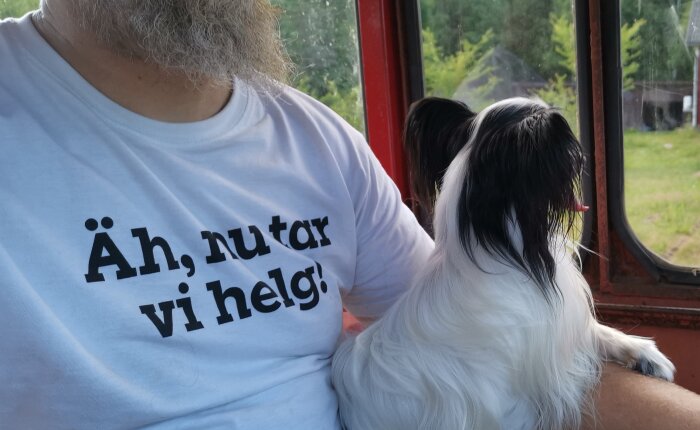 Man i vit t-shirt och hund tittar ut genom fönster. Text på tröja: "Åh, nu tar vi helg!"