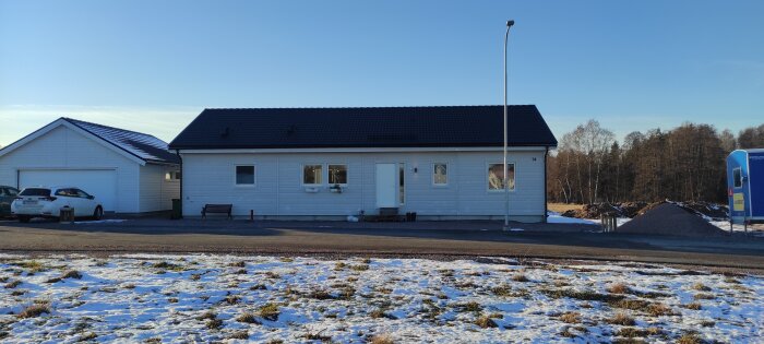 Enfamiljshus med snöfläckar, garage, lantlig omgivning, blå himmel, något byggmaterial i bakgrunden.