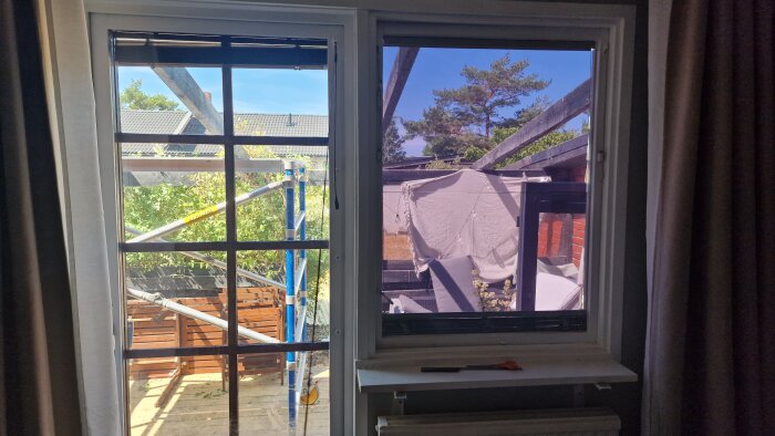 Utsikt genom fönster på byggnadsställning, hängande tvätt, trädgård och blå himmel.