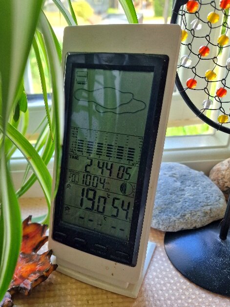 Digital väderstation inne med tid, datum, temperatur, avfuktighet. Fönster, växt, stenar och dekorativ lampa syns också.