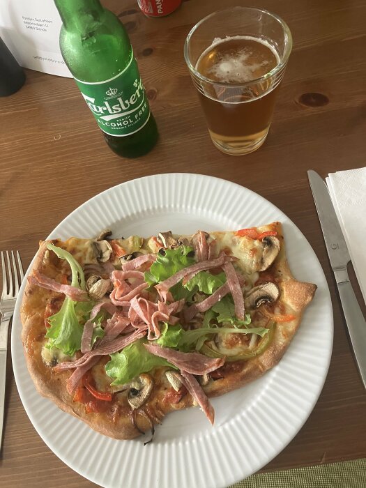 Pizza med skinka, sallad, svamp på vit tallrik, alkoholfri öl Carlsberg och glas bredvid.