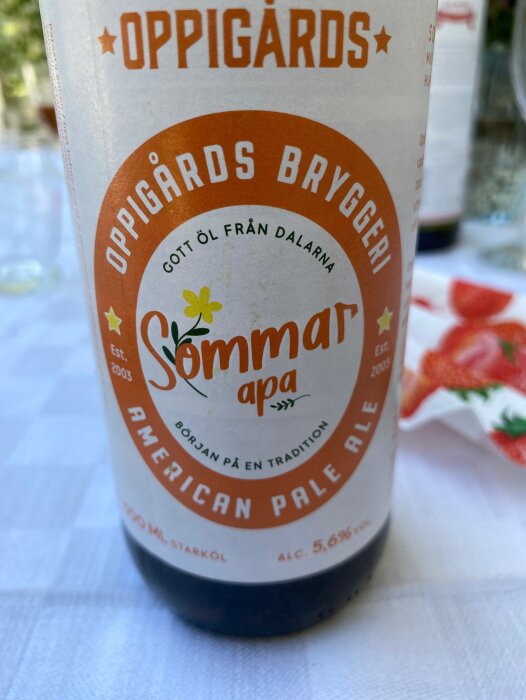 En ölflaskeetikett: Oppigårds Bryggeri, Sommar APA, American Pale Ale, 5.6% alkohol, vitt bord i bakgrunden.