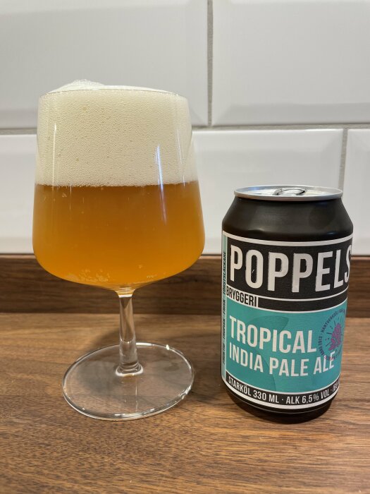 Ett glas med skummande öl och en burk Poppels Tropical India Pale Ale på en träyta.