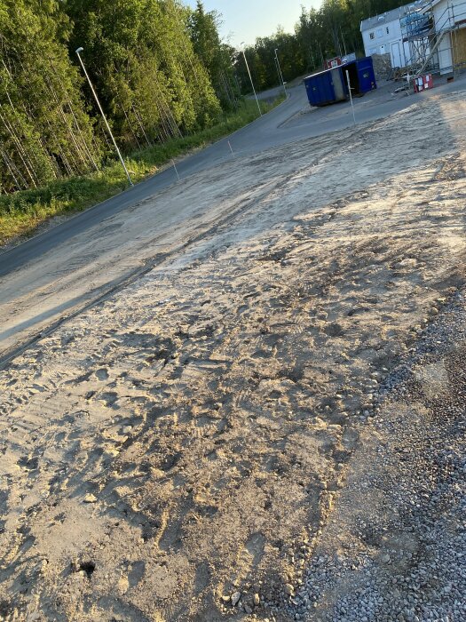 Industriområde, grusväg, spår efter fordon, byggnadsställning, containrar, gröna träd, skylt, kvällssol.