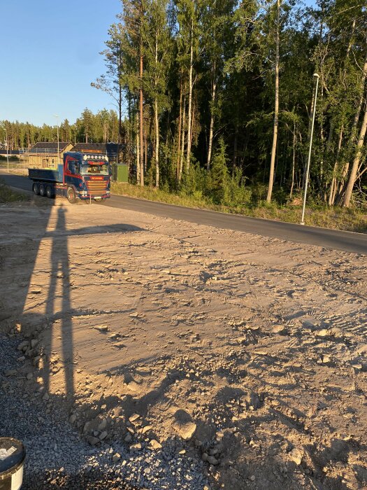 Lastbil på grusväg nära skog, byggnadsarbete, lång skugga, solljus, klar himmel, sommar eller vår.