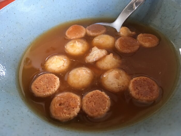 En skål med klarbrun buljong och flytande små rundbröd, troligtvis köttbuljong med pannkaksskivor eller croutoner.