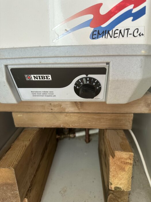 NIBE värmepanna med EMINENT-Cu logotyp, temperaturvred, varningsinformation, upphöjd på träreglar.