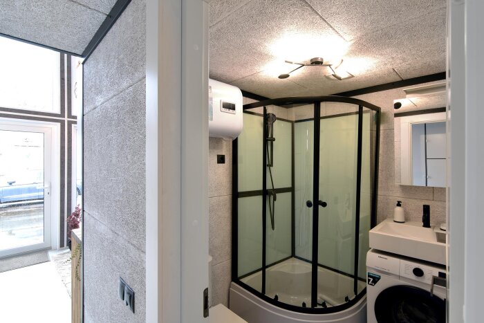 Modernt badrum med duschkabin, handfat, spegel och tvättmaskin. Ljus inredning och fönster.