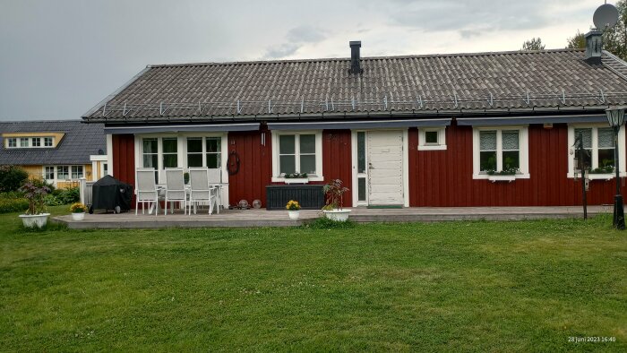 Rött trähus med vita knutar, gräsmatta, utemöbler på trädäck, molnig himmel, sommardag.