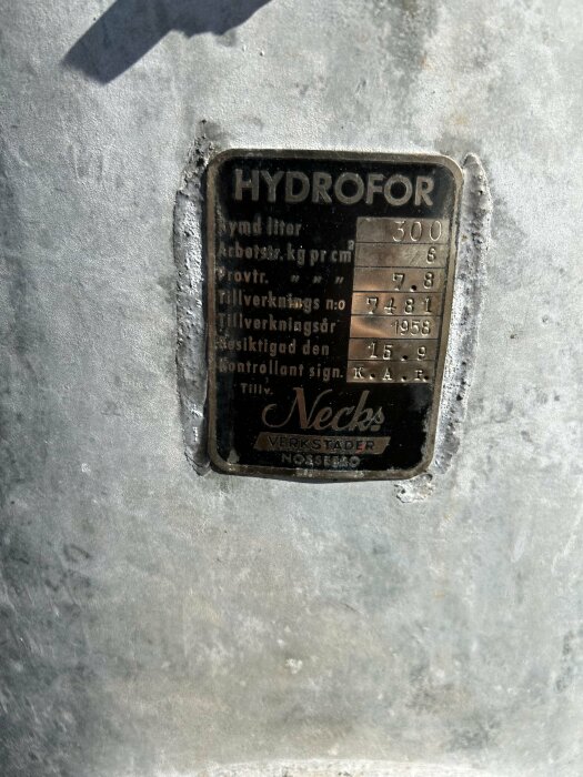 Metallskylt för hydrofor med tekniska specifikationer; solblekt, slitet, på grov yta.