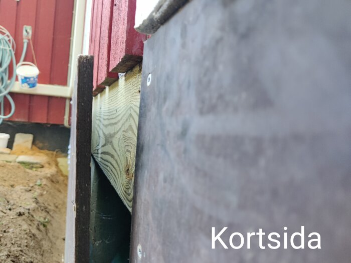 Delvis suddig bild, trästrukturer, röd vägg, blå hink, vit ledning, märkt med "Kortisda". Utomhus, byggnadsdetaljer, otydligt fokus.