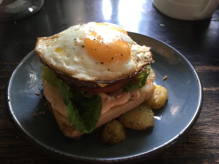 Stekt ägg på BLT-sandwich med rostade potatisar på blå tallrik, träbord.