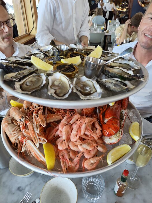 Skaldjurstorn med ostron och räkor, personer vid restaurangbord, festlig atmosfär.