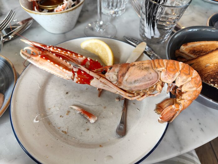 Halväten hummer på tallrik, citronskiva, rostat bröd, skaldjursrester, matplats med bestick och glas.