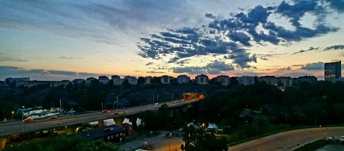 Stadslandskap vid skymning, vägar, bilar, byggnader och dramatiskt molnigt himmel.