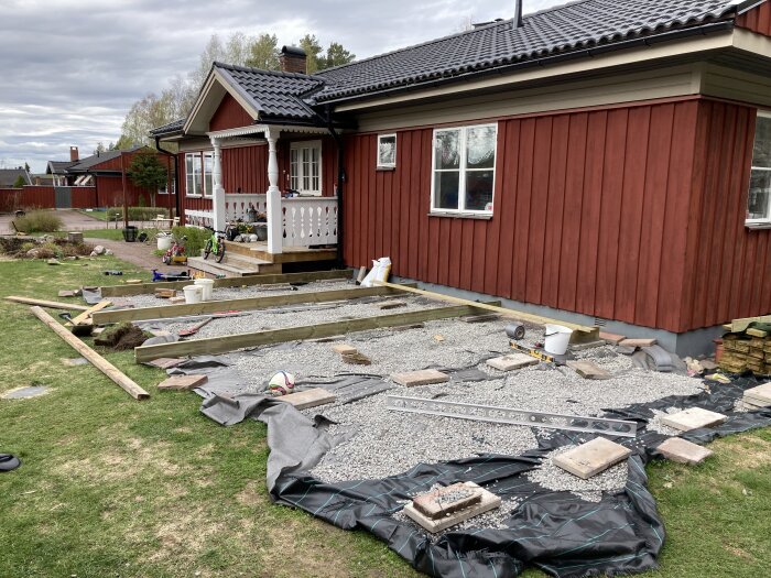 Rött hus under renovering, altanbygge pågår, byggmaterial och verktyg synliga på gräsmattan.