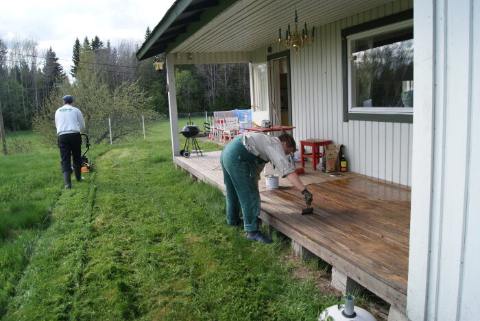 Två personer arbetar utomhus, en klipper gräsmatta och den andra målar veranda.