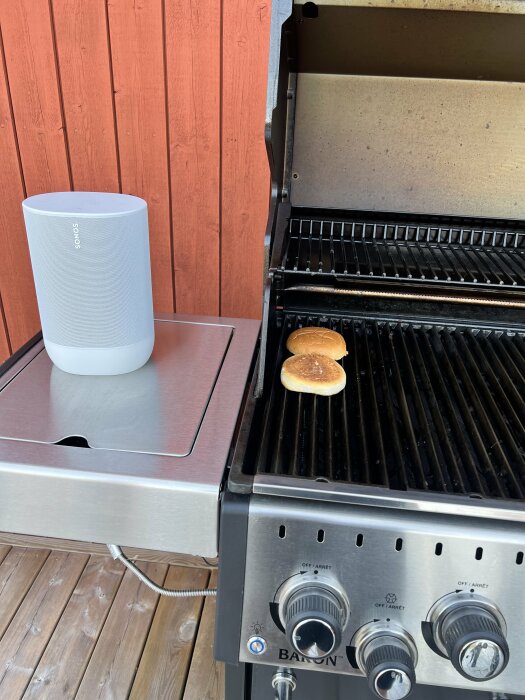 Trådlös högtalare på grillbord, ett uppklippt hamburgarbröd på stängd gasolgrill, träbakgrund.