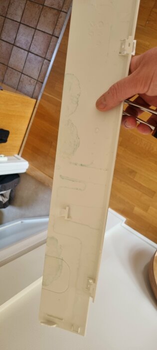 En person håller en smutsig vit plastdel mot inomhusbakgrund med kakel- och trägolv.