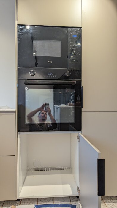 Kök med inbyggd mikrovågsugn och ugn, spegelbild av fotografen i glaset, skåplucka öppen.