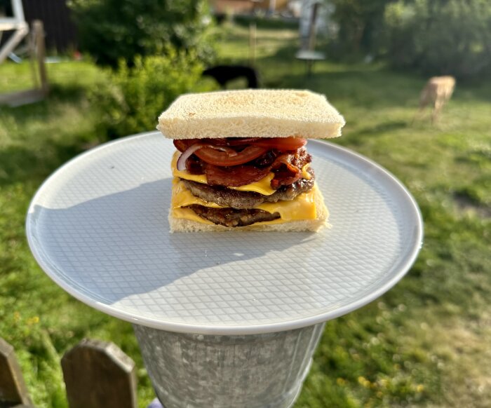 Hamburgare med ost, bacon och tomat på vitt bröd, utomhus på en tallrik. Bakgrunden visar en trädgård.