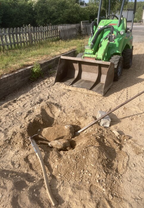 Grön minilastare, skopa nära grävd grop, kratta, spett på sandig mark, staket i bakgrunden.