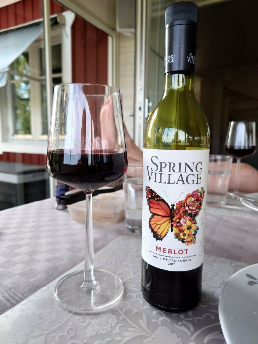 Ett glas rött vin och en flaska Merlot på ett bord utomhus med en suddig bakgrund.
