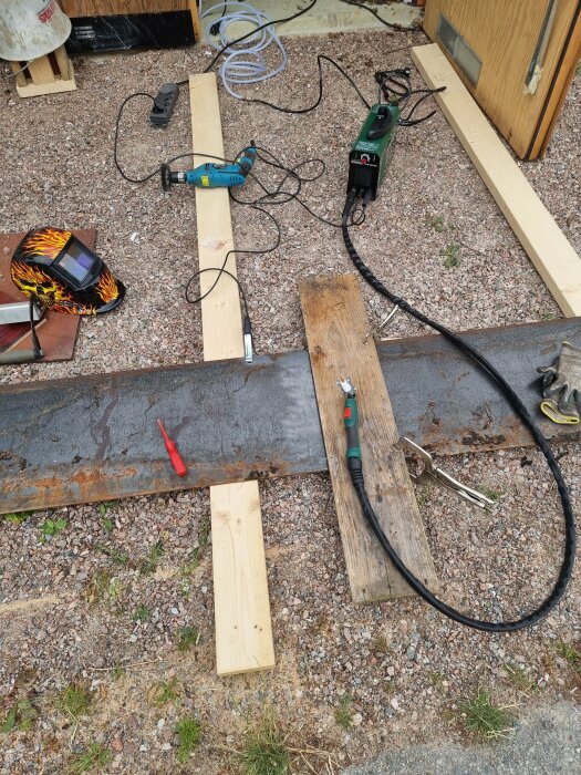 Verktyg och material på marken, byggarbetsplats, svetshjälm med lågflammigt mönster, elverktyg, träplankor, kablar.