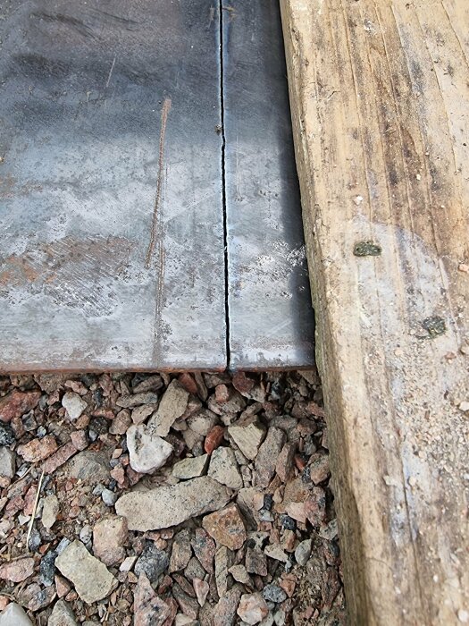 Stålyta möter träplanka ovanpå grus och småstenar. Slitna material. Ojämna ytor. Kontrast mellan natur och industri.