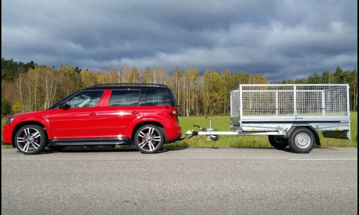 Röd SUV med släpvagn parkerad vid sidan av en väg, molnig himmel, grön skog i bakgrunden.
