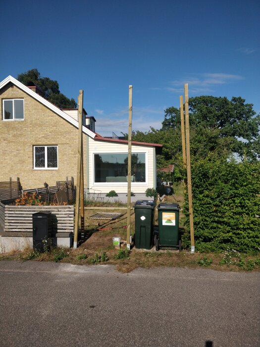 Ett bostadshus, höga stolpar i trädgården, grönska, soptunnor framför, soligt väder.