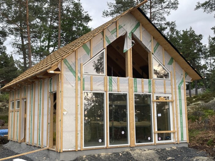 Ett hus under konstruktion med trästomme, isoleringsmaterial, stora fönster och omgiven av natur.