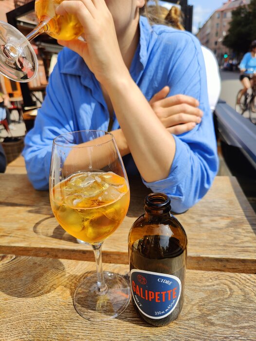 Person dricker cider utomhus vid träbord, Galipette-flaska, isbitar och citron i glaset.