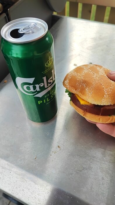 En hand håller en hamburgare, bredvid en öppnad Carlsberg ölburk, utomhus på solig dag.