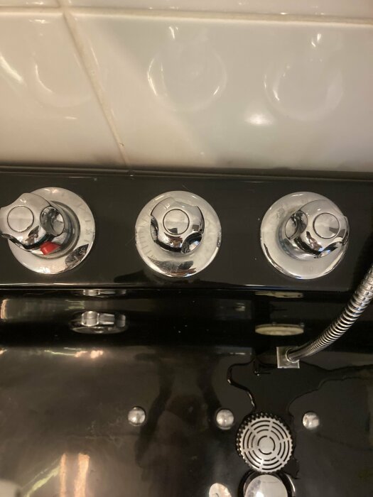 Svart spis med tre kontrollvred och ett kokplattsmönster, mot en kaklad vägg.