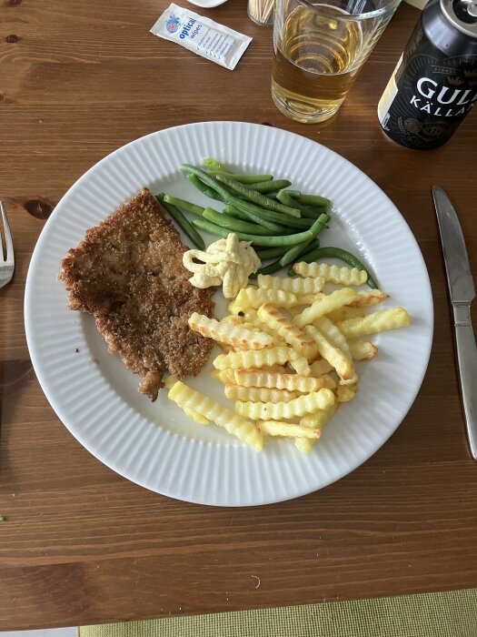 Tallrik med schnitzel, pommes, gröna bönor och bearnaisesås, ölburk och majonnispåse i bakgrunden.