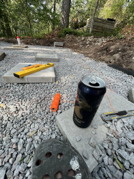 Byggarbetsplats med nivelleringverktyg, ölburk, grus och betongplattor, bland verktyg under konstruktion.