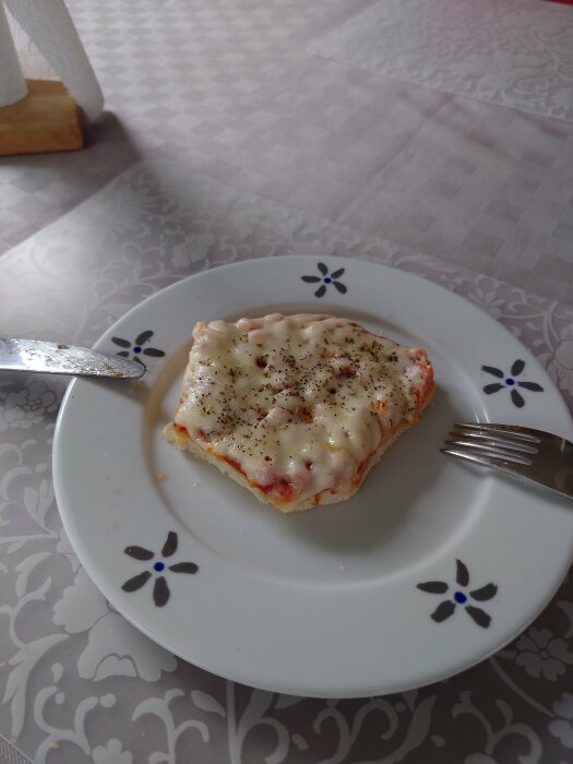 En gratinerad maträtt på tallrik, troligen lasagne, med oregano ovanpå. Bestick till vänster. Blommigt bord och servetthållare.